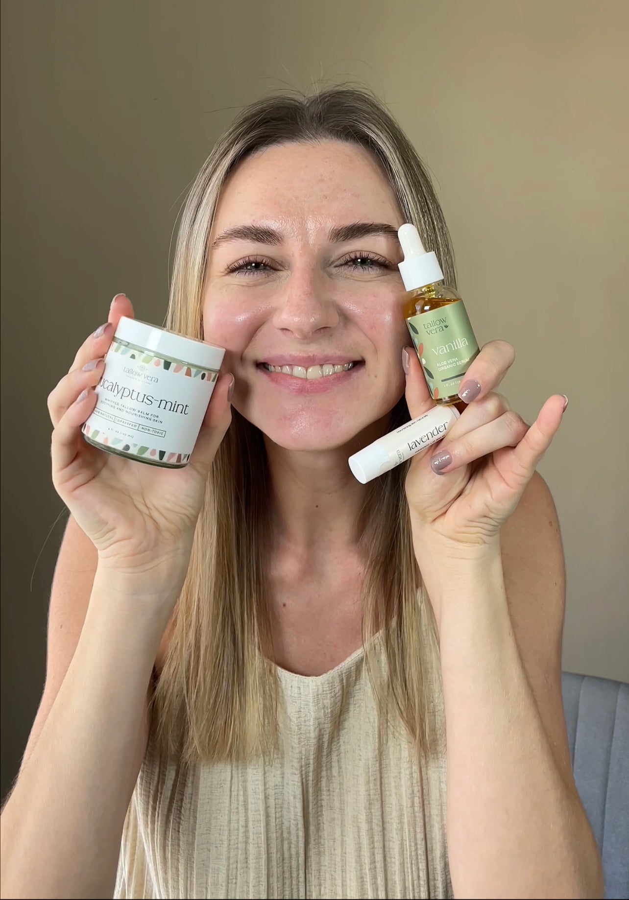 Woman holding a 4oz jar of tallow, tallow lip balm and bottle of aloe vera skincare products against a neutral background
