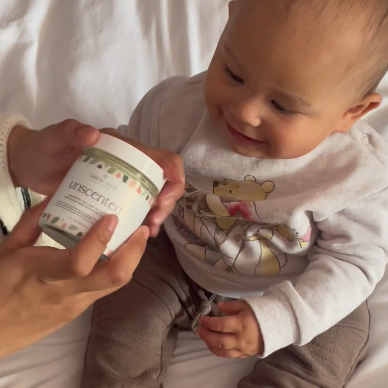 Baby sitting on a white surface with a person holding a container labeled 'TALLOW VERA'.