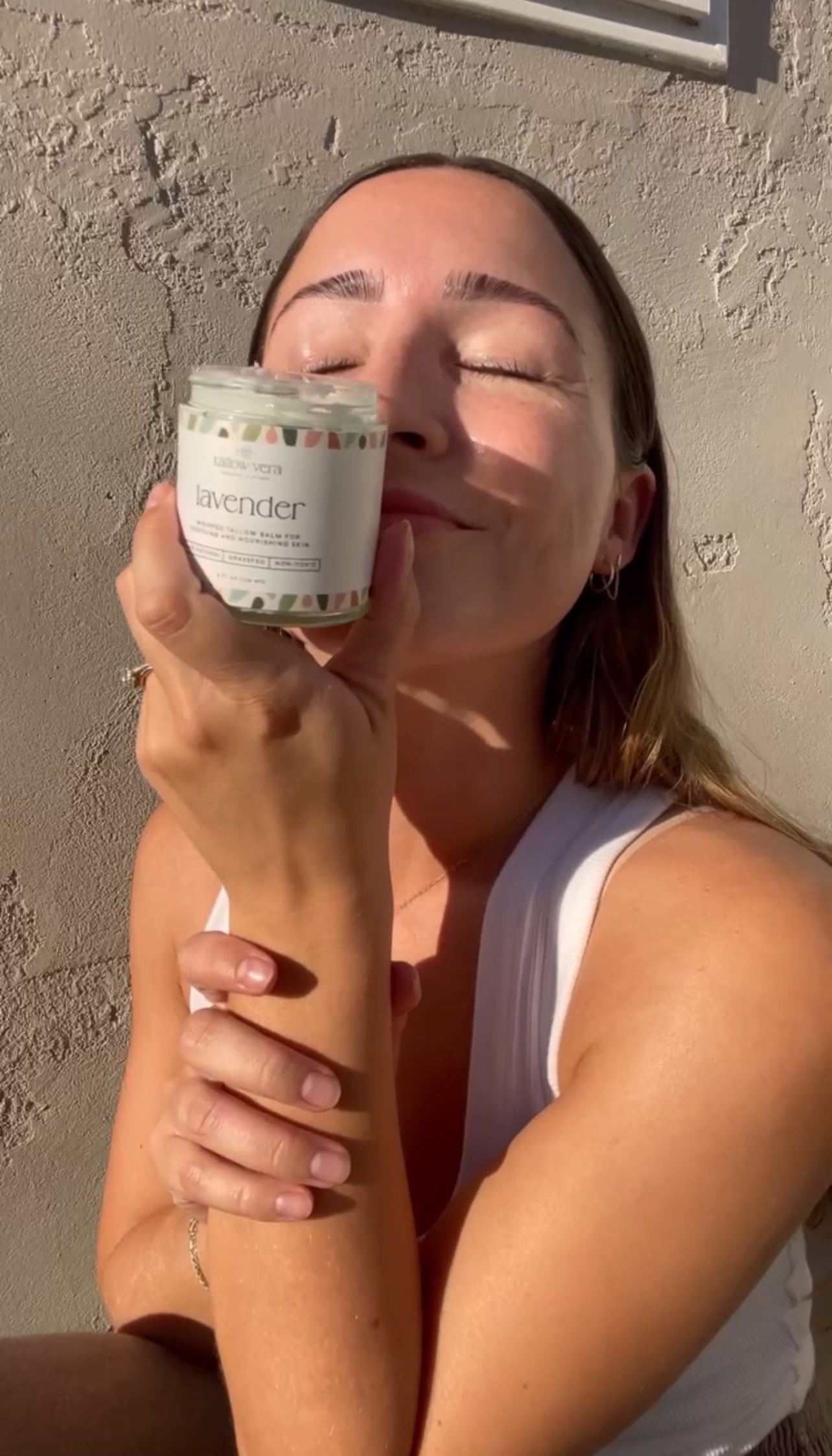 Woman holding a 4oz Tallow Vera tallow balm labeled 'Lavender' against a textured wall outside in the sun.