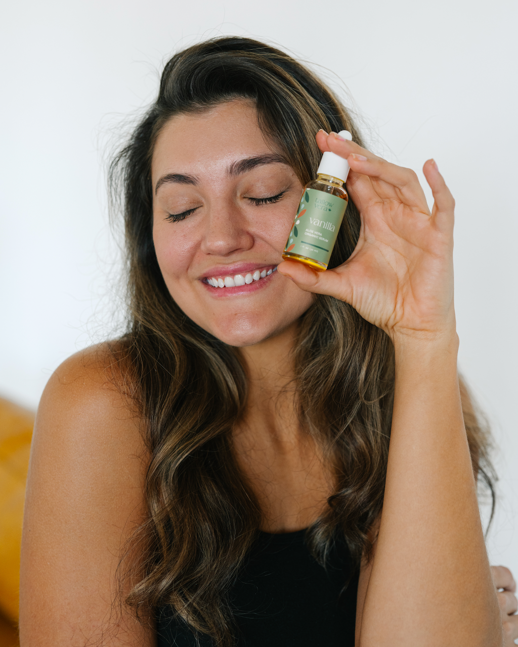 Woman holding a bottle of vanilla Tallow Vera serum near her face against a white background